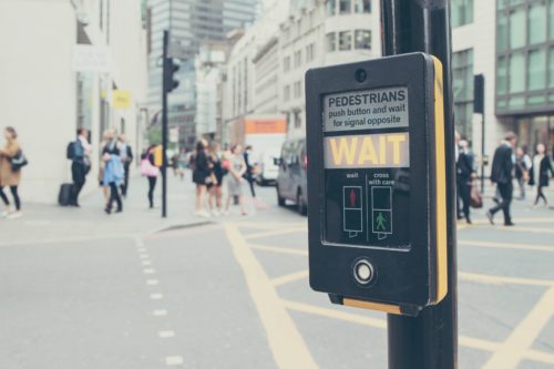 A pedestrian crossing button in an urban area, where people wait to cross safely amid concerns about pedestrian accidents. The button reads "WAIT" and instructs users to push for the signal opposite. Buildings and vehicles form the busy backdrop.