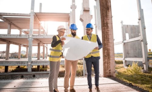 Three people in safety gear, including helmets and vests, carefully examine a large blueprint at a construction site. With the unfinished building structure looming in the background under a clear sky, they discuss designs that ensure no future sidewalk accidents occur.