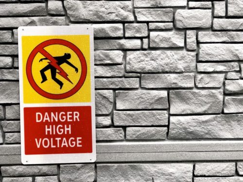 A "Danger High Voltage" warning sign depicting electrocution with an image of a person being struck by lightning is mounted on a stone wall. The background features textured grey bricks.